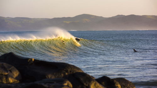 A surfer riding a wave at Manu Bay, Raglan, with rocky headland in the foreground