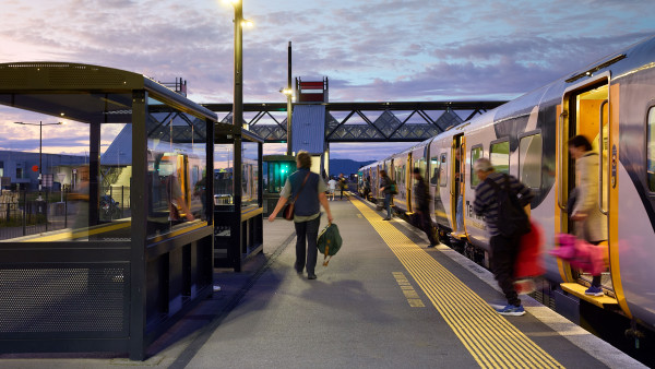 Te Huia train stopped at Rotokauri station looking north, with overbridge in distance and people getting out of carriages.